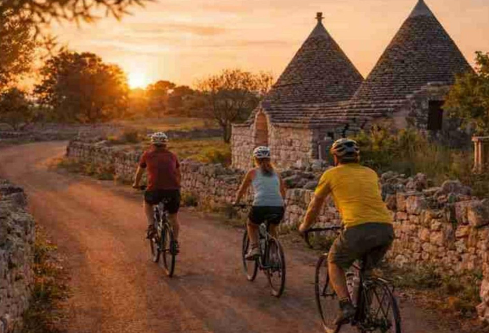 Cyclists on a gravel road at sunset, passing trulli and dry-stone walls in Salento